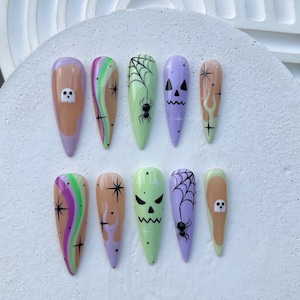 May include: A set of ten long, almond-shaped press-on nails with Halloween-themed designs. The nails feature a variety of colors, including purple, green, and orange, with designs such as ghosts, spiderwebs, and flames. The nails are arranged on a white surface.