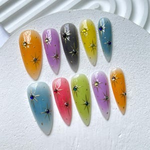 Puede incluir: Conjunto de diez u&ntilde;as postizas largas y ovaladas con un degradado de color de claro a oscuro. Cada u&ntilde;a tiene un dise&ntilde;o de estrella dorada y un solo strass.