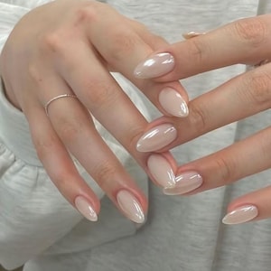 May include: Almond-shaped fingernails with a glossy, light beige, semi-sheer finish. The nails feature a smooth, reflective surface and a clean, rounded tip. A thin, simple metallic ring is worn on the ring finger, complementing the neutral manicure.