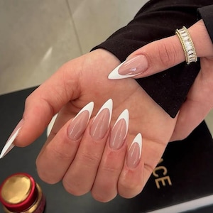 Pode incluir: Close-up de uma mão com unhas longas e em forma de amêndoa pintadas com uma manicure francesa clássica. As unhas são de cor rosa claro com uma ponta branca.