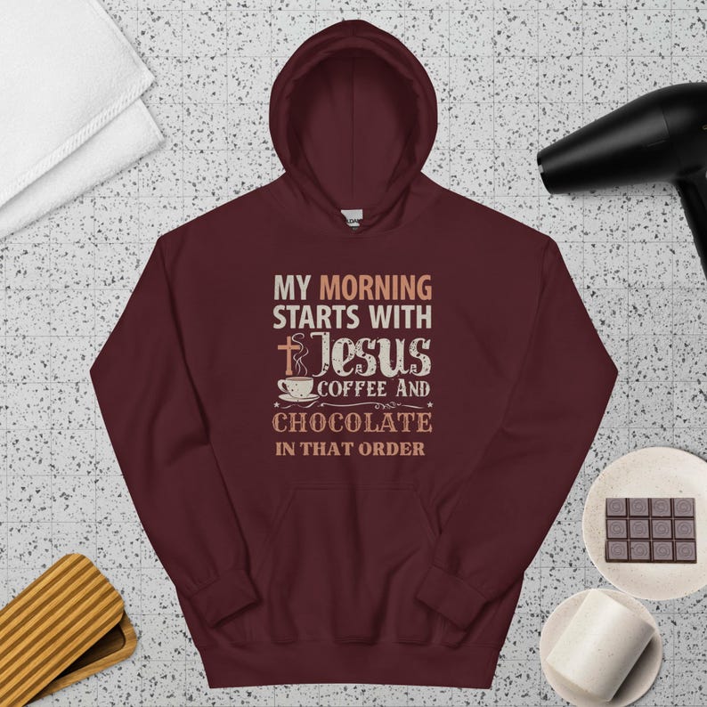 Peut inclure: Un sweat &agrave; capuche bordeaux avec le texte "MY MORNING STARTS WITH JESUS COFFEE AND CHOCOLATE IN THAT ORDER". Le sweat &agrave; capuche a une capuche &agrave; cordon et une poche avant. Une serviette blanche, un s&egrave;che-cheveux et une barre chocolat&eacute;e sont en arri&egrave;re-plan.