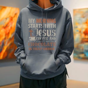 Peut inclure: Un sweat-shirt bleu ardoise avec une poche avant et une capuche &agrave; cordon. Le devant du sweat-shirt affiche le texte "MY MORNING STARTS WITH JESUS COFFEE AND CHOCOLATE IN THAT ORDER" en blanc et orange, avec une croix et une tasse de caf&eacute;.