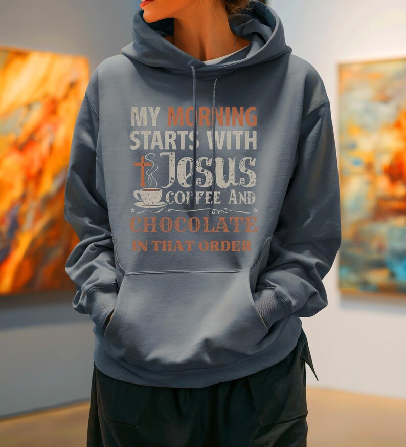Peut inclure: Un sweat-shirt bleu ardoise avec une poche avant et une capuche &agrave; cordon. Le devant du sweat-shirt affiche le texte "MY MORNING STARTS WITH JESUS COFFEE AND CHOCOLATE IN THAT ORDER" en blanc et orange, avec une croix et une tasse de caf&eacute;.