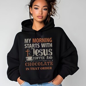 Peut inclure: Un sweat &agrave; capuche noir avec l'inscription "MY MORNING STARTS WITH Jesus COFFEE AND CHOCOLATE IN THAT ORDER" en blanc, orange et marron. Le motif comprend une croix, une tasse &agrave; caf&eacute; et des &eacute;toiles.