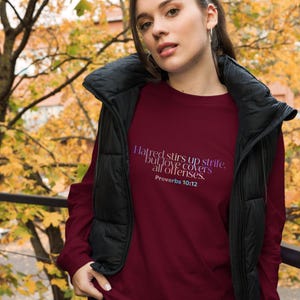 Puede incluir: Una camiseta de manga larga color burdeos con una cita en degradado de colores morado y azul que dice "El odio provoca contiendas, pero el amor cubre todas las ofensas. Proverbios 10:12"