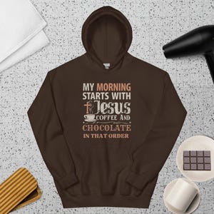 Peut inclure: Un sweat-shirt &agrave; capuche marron avec le texte "MY MORNING STARTS WITH Jesus COFFEE AND CHOCOLATE IN THAT ORDER." Le design comprend une croix, une tasse &agrave; caf&eacute; et des &eacute;toiles. Le sweat-shirt est pr&eacute;sent&eacute; sur une surface &agrave; motifs.