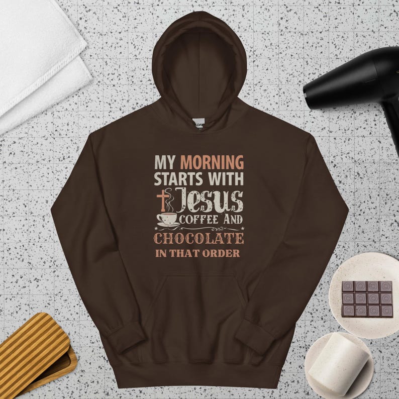 Peut inclure: Un sweat-shirt &agrave; capuche marron avec le texte "MY MORNING STARTS WITH Jesus COFFEE AND CHOCOLATE IN THAT ORDER." Le design comprend une croix, une tasse &agrave; caf&eacute; et des &eacute;toiles. Le sweat-shirt est pr&eacute;sent&eacute; sur une surface &agrave; motifs.