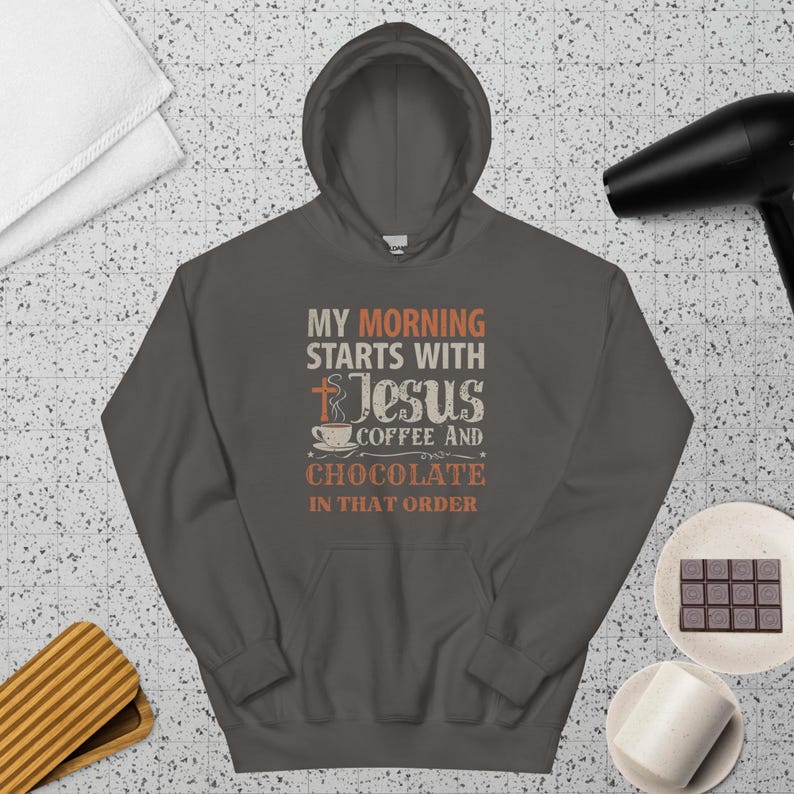 Peut inclure: Un sweat &agrave; capuche gris fonc&eacute; avec l'inscription "MY MORNING STARTS WITH JESUS COFFEE AND CHOCOLATE IN THAT ORDER". Le design comprend une croix, une tasse &agrave; caf&eacute; et une barre de chocolat.