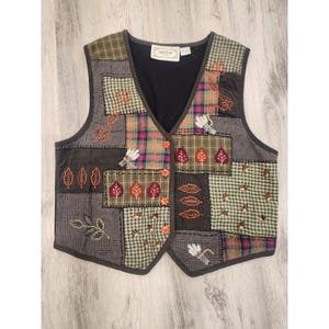 Puede incluir: Un chaleco patchwork con cuello en V. El chaleco presenta varios patrones de tela, incluyendo cuadros y colores lisos, con diseños bordados de hojas y trigo. Tiene botones naranjas y una espalda negra.