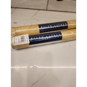 May include: Two rolled wallpaper rolls with a beige pattern. Each roll has a navy blue band with the text "RALPH LAUREN HOME". One roll has a label with text and a barcode.