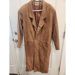 Può includere: Un lungo cappotto in camoscio marrone con collo a rever e due tasche frontali. Il cappotto ha una chiusura con bottoni ed è appeso a una gruccia bianca. Il marchio "FORENZA" è visibile sull'etichetta.