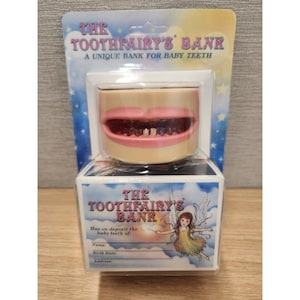 May include: A plastic container shaped like a mouth with pink lips and white teeth, labeled The Toothfairy's Bank. The packaging features a blue sky background with stars, an illustration of a fairy, and blank lines for name, birth date, and address.