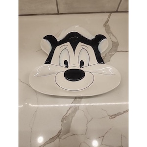 May include: A white ceramic dish shaped like a cartoon character's face, featuring black and white details. The dish has a rounded shape with a large black nose, eyes, and a black outline.
