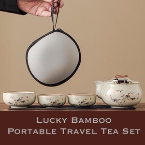 Puede incluir: Un juego de té portátil crema con una tetera, tres tazas y un estuche de transporte. Las piezas de cerámica presentan diseños de ramas de bambú. El juego incluye una alfombrilla negra y se exhibe sobre una superficie de madera.