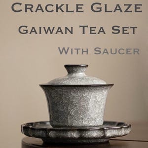 Könnte beinhalten: Ein graues Gaiwan-Teeservice mit Craquelé-Glasur und Untertasse. Das Set enthält eine Tasse mit Deckel und eine passende Untertasse. Tasse und Untertasse haben eine strukturierte Oberfläche. Der Text "CRACKLE GLAZE GAIWAN TEA SET WITH SAUCER" ist zu sehen.