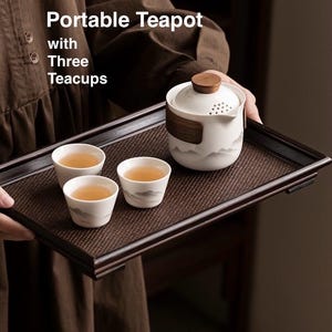 Peut inclure: Un service à thé portable blanc avec un couvercle et des accents en bois, accompagné de trois tasses assorties, le tout présenté sur un plateau marron foncé. Les tasses et la théière présentent un motif de montagne minimaliste. Le texte "Portable Teapot with Three Teacups" est visible.