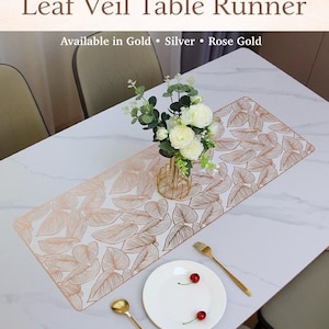 May include: A rose gold leaf-patterned table runner on a white table, with a vase of flowers, a plate with cherries, and gold cutlery. The text "Leaf Veil Table Runner" is at the top, with "Available in Gold • Silver • Rose Gold" below.