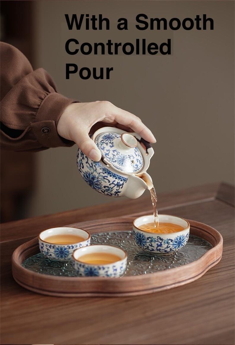 May include: A small, ornate teapot with a blue and white floral design is pouring tea into matching cups. The cups and teapot sit on a wooden tray with a glass insert. The text "With a Smooth Controlled Pour" is at the top of the image.