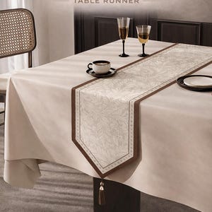 May include: A beige table runner with a leaf pattern and brown trim, displayed on a white tablecloth. The text "Vintage Leaf Veil TABLE RUNNER" is at the top. A coffee cup, two wine glasses, and a plate are on the table.