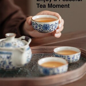May include: A hand holding a small teacup filled with tea, with a blue and white floral design. The image includes a teapot and two other teacups on a tray. Text reads: "Enjoy a Peaceful Tea Moment."