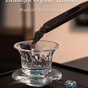 May include: A clear crystal teacup and saucer set with a decorative design. Tea leaves are being poured into the cup from a wooden scoop. The image text reads "Beautiful Crystal Teaware".