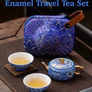 Puede incluir: Un juego de té de viaje de esmalte azul y blanco con el texto "Hundred Blossoms Enamel Travel Tea Set". El juego incluye una tetera, dos tazas de té y un estuche de transporte con un asa de cuero. La tetera y las tazas tienen un diseño floral.
