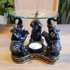 May include: A black and silver elephant-themed oil burner featuring three sculpted elephants supporting a clear glass dish. The base is decorated with gold trim and includes a central circular holder for a small tea light candle.