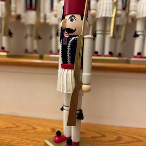 May include: A wooden nutcracker soldier in a traditional uniform. The nutcracker has a red hat, a blue jacket with gold trim, and a white pleated skirt. It holds a gold-colored rifle and stands on a gold base with red shoes.