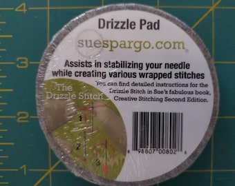 May include: A round, gray Drizzle Pad for needlework, wrapped in clear plastic. The label reads "Drizzle Pad" and "suespargo.com." Text on the label describes the product's use in stabilizing needles for wrapped stitches. Includes a diagram and a barcode.