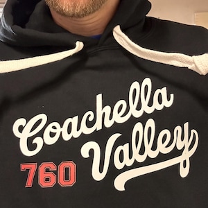 Può includere: Felpa con cappuccio nera con coulisse bianca e le parole "Coachella Valley" in un font corsivo. Il numero "760" è in rosso, in un font a blocchi. La felpa è realizzata in un materiale morbido.