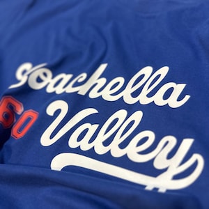 Puede incluir: Camiseta azul real con las palabras "Coachella Valley" en escritura blanca. El número "60" es rojo con un contorno blanco. La camiseta está hecha de un material suave.