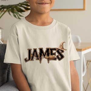 May include: A cream-colored t-shirt featuring the name "JAMES" in a black and gold font. A brown wizard hat and broom are incorporated into the design. The shirt is made of a soft material.