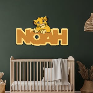 May include: A wooden wall decoration featuring the name "Noah" in large, yellow block letters. A cartoon lion cub is perched above the name. The decor is set against a dark green wall, with a crib visible below.