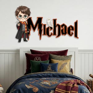 May include: A wall decal featuring a cartoon depiction of a character with glasses, a wand, and a scarf, alongside the name "Michael" in a stylized font. A white owl sits atop the name. The decal is above a bed with decorative pillows and a patterned comforter.