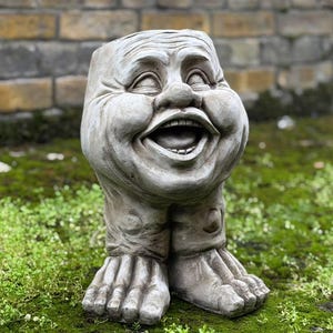 以下が含まれることがあります： 足と足で支えられた、歯が見える笑顔の顔を描いた、気まぐれな灰色のコンクリート製の庭の彫像。彫像は、背景にレンガの壁がある草地エリアに設置されています。