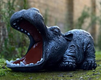 Puede incluir: Una escultura de cerámica negra de un hipopótamo con la boca abierta de par en par, mostrando sus dientes. La escultura está tumbada de lado sobre un lecho de musgo verde.