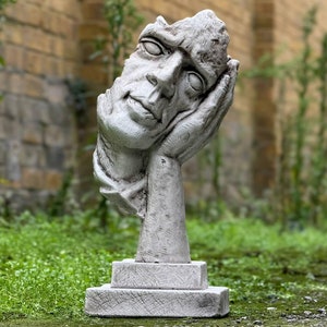 Pode incluir: Uma escultura de concreto branco de uma cabeça humana com uma mão apoiada no queixo. A escultura está sobre uma base quadrada e é colocada sobre um fundo de grama verde.