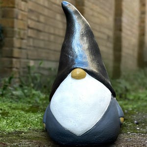 Puede incluir: Estatua de gnomo de jardín con un gorro puntiagudo negro, barba blanca y traje gris. El gnomo tiene una nariz pequeña y redonda de color dorado. La estatua está hecha de un material similar a la piedra y se encuentra sobre hierba verde y un muro de ladrillos.