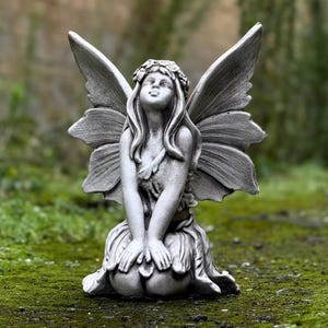 Puede incluir: Una escultura de piedra gris de un hada con alas, sentada con las manos juntas en su regazo. El hada lleva una corona de flores y mira hacia arriba.