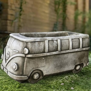 May include: A gray concrete planter shaped like a vintage van. The planter has detailed features including headlights, wheels, and a front emblem. It is designed to hold plants and sits on a bed of green moss.