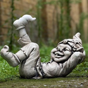 Puede incluir: Una estatua de gnomo de jardín de piedra gris tumbada de espaldas con una pierna levantada en el aire. El gnomo tiene una expresión caprichosa en la cara y lleva un sombrero.