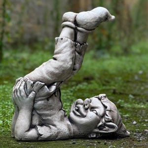 Puede incluir: Una estatua de piedra gris de un gnomo haciendo una parada de manos. El gnomo lleva pantalones y tiene una expresión traviesa en su rostro.