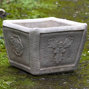 Könnte beinhalten: Ein quadratischer, grauer Betontopf mit aufwendigen Blumen- und Blattmotiven. Der Topf hat eine raue, strukturierte Oberfläche.