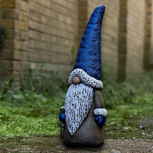Puede incluir: Una estatua de gnomo de jardín con un sombrero azul largo y puntiagudo y una larga barba blanca. El gnomo lleva un abrigo marrón con ribetes blancos y guantes azules. La estatua está sobre una superficie de piedra.
