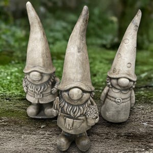 Puede incluir: Tres gnomos de jardín de piedra con sombreros altos y puntiagudos. Los gnomos tienen caras sonrientes, largas barbas y visten trajes con delantales. Uno de ellos sostiene una paleta y una cesta de flores.