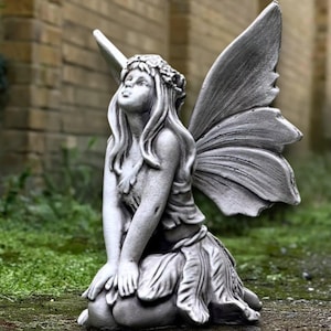 May include: A gray stone fairy statue kneeling with large, detailed wings. The fairy wears a floral crown and a leaf-like skirt. The statue is set on a green, grassy surface with a brick wall in the background.