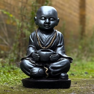 Puede incluir: Estatua negra de un monje meditando sentado, con los ojos cerrados, sosteniendo un cuenco. El monje lleva una túnica y un collar de cuentas. La estatua está sobre una base redonda y está al aire libre.
