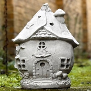 Puede incluir: Una casa de hadas de hormigón gris con un techo de hongo, una puerta y ventanas. La casa tiene la palabra "Home" escrita en la parte delantera.