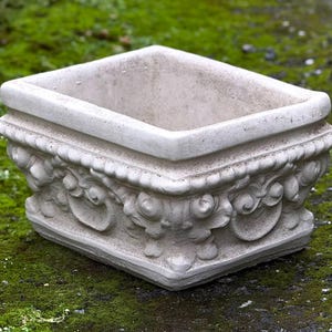 May include: A white rectangular planter with ornate detailing around the top edge. The planter is made of a textured material and has a slightly weathered look.