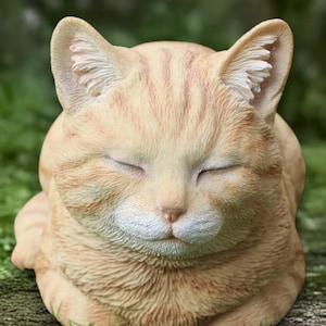 May include: A light orange cat ornament with closed eyes, depicting a peaceful expression. The cat's fur has delicate, darker orange stripes. The sculpture is set against a blurred green background, suggesting an outdoor setting.